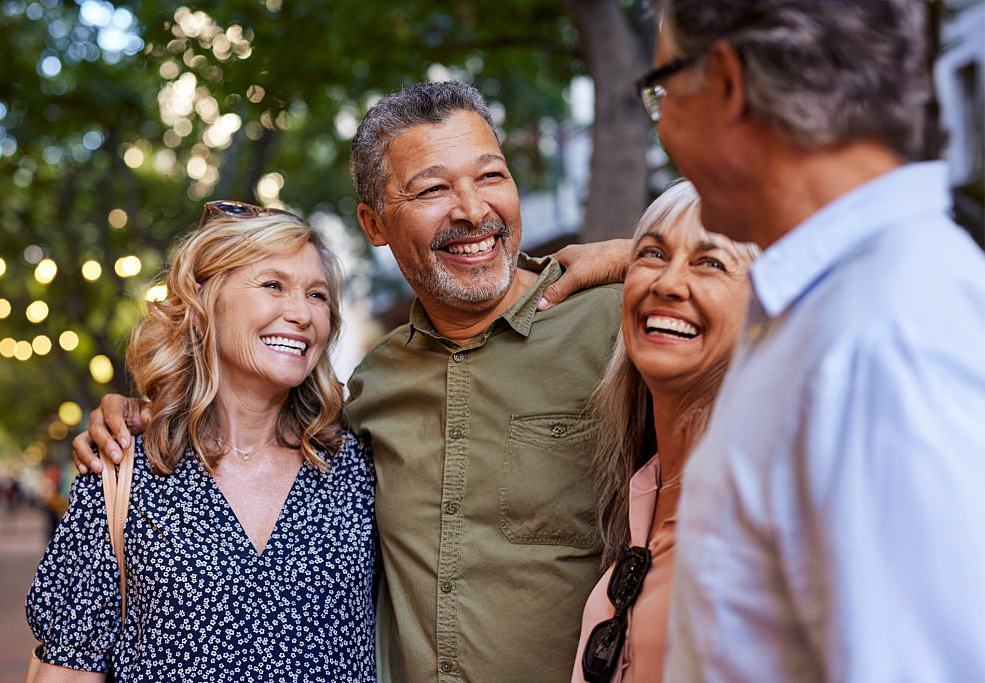 Four middle-aged adults on a street at dusk, standing close and smiling, one man with arms around two women.