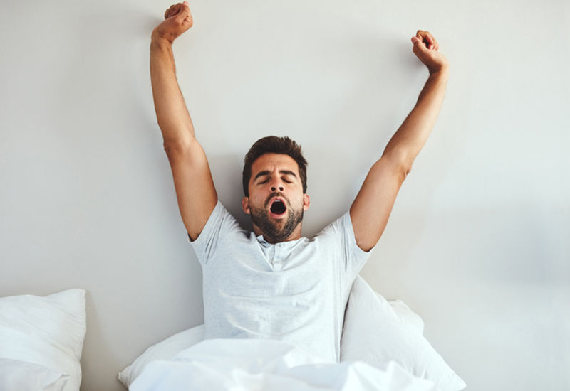 Adult man sitting up in bed, yawning and stretching his arms overhead against white bedding and a light gray wall.
