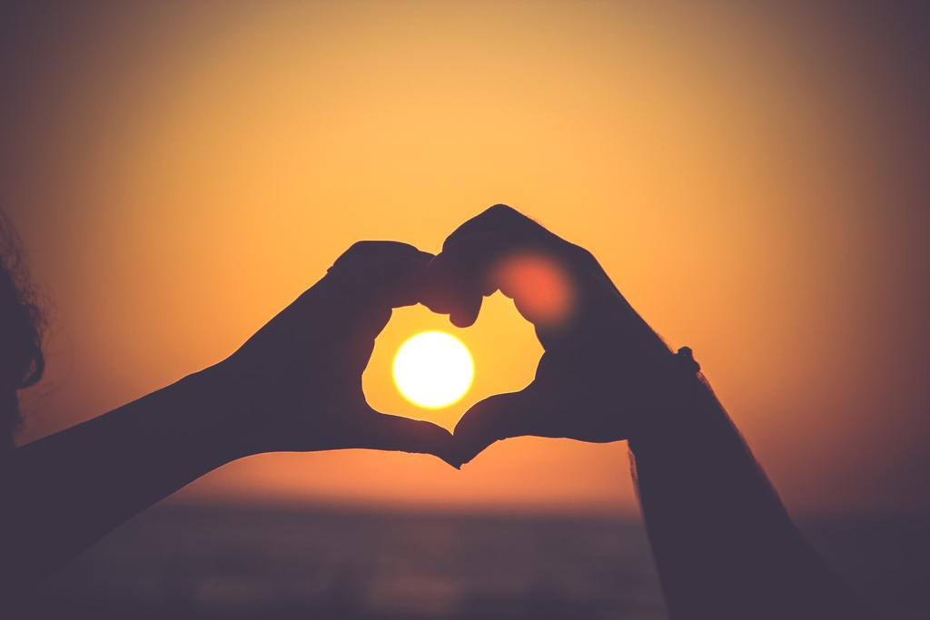 Two silhouetted hands forming a heart framing the sun against an orange-yellow sky over a blurred water horizon.