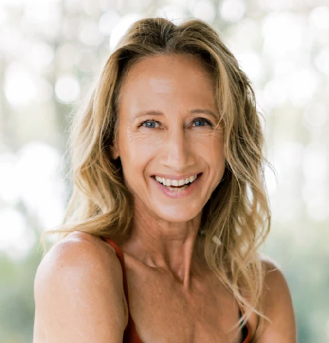 Close-up portrait of an adult woman with wavy blonde hair smiling in a rust sleeveless top against soft green bokeh.