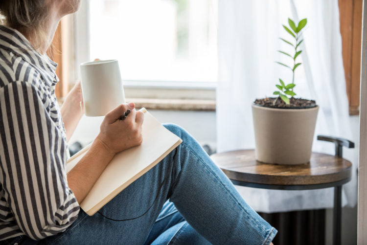 Person in black-and-white striped shirt sits by a window, holding a white mug and pen with a notebook on their knee.