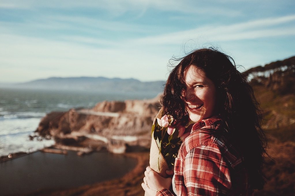 Young woman in a red plaid shirt smiling over her shoulder while holding a bouquet of pink tulips by a rocky coastline.