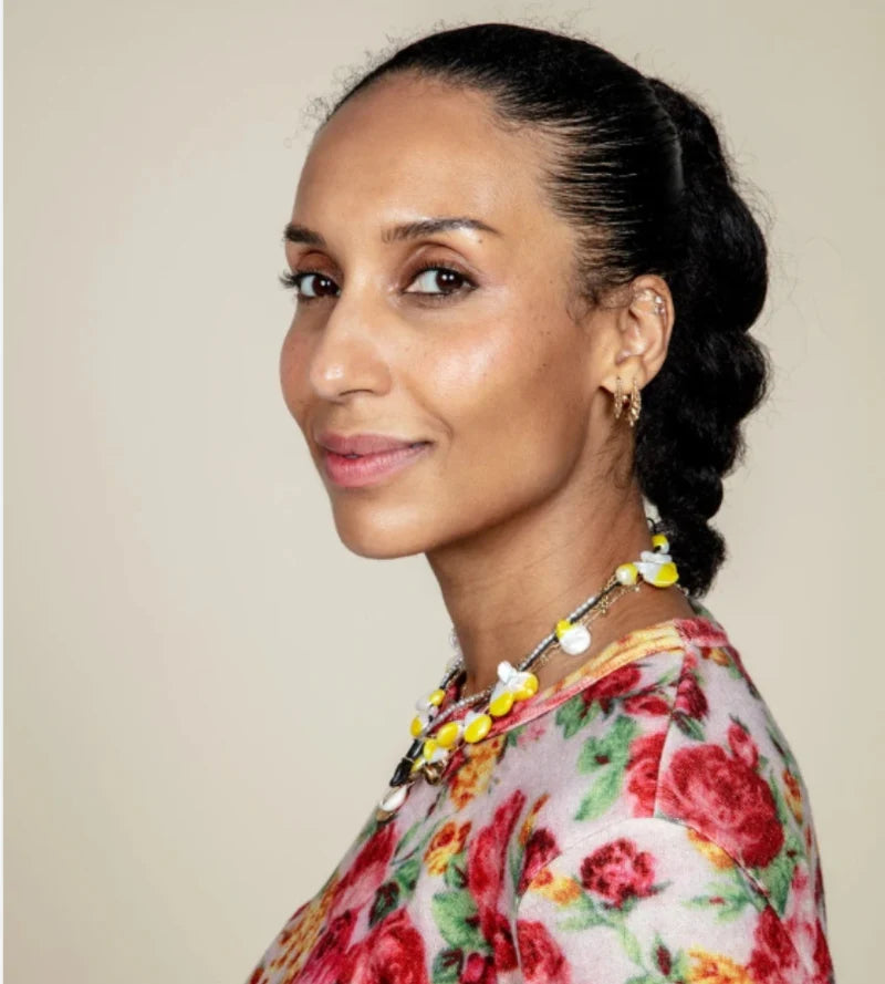 Woman with braided hair wearing a floral top and a yellow necklace against a plain background