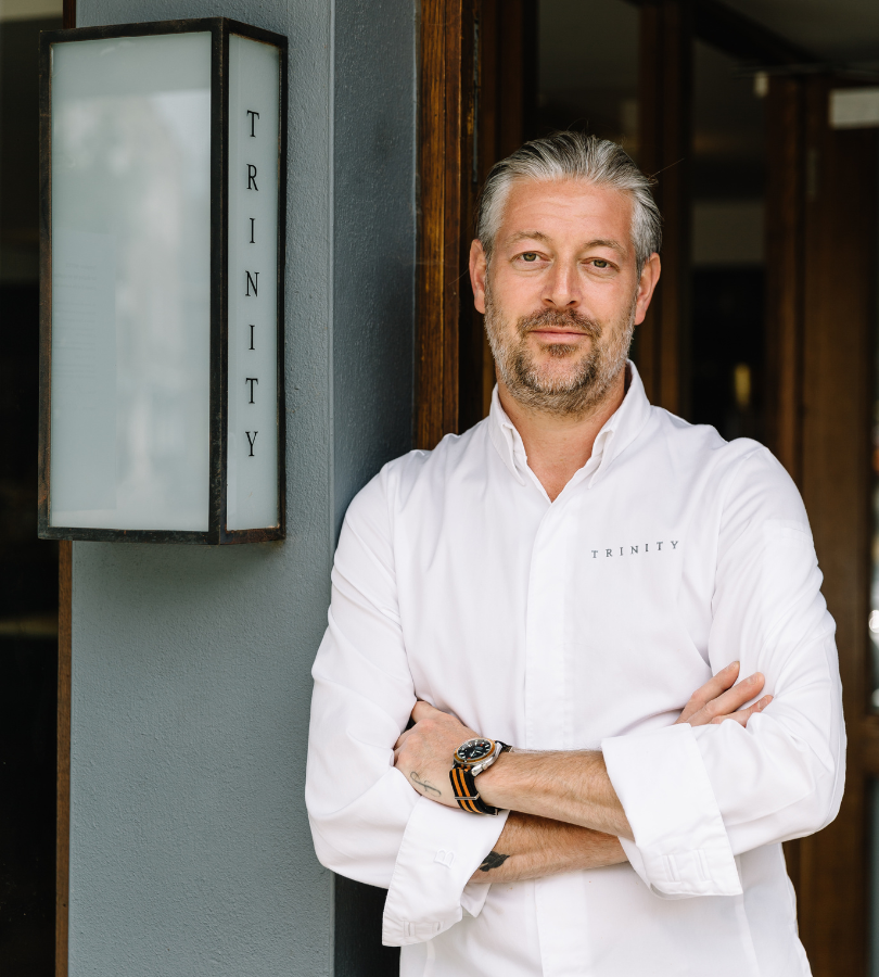 Middle-aged man with grey hair and beard wearing a white chef coat standing with arms crossed by a wall sign reading Trinity