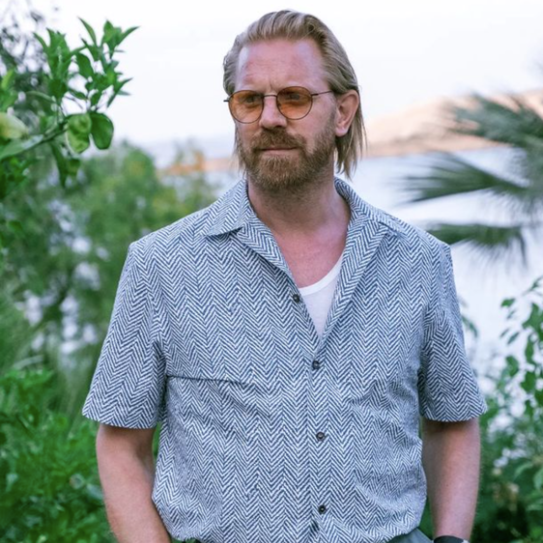 Man with beard and glasses wearing patterned short-sleeve shirt standing outdoors near greenery in soft daylight