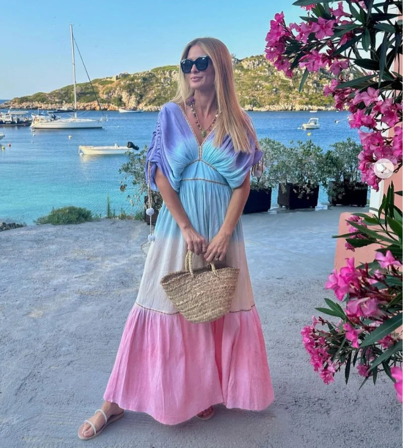 Woman in a colorful dress, carrying a bag and wearing sunglasses, standing by a scenic waterfront with boats and flowers.