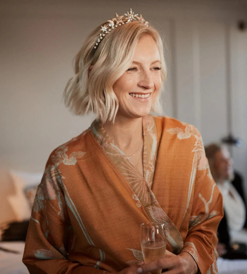 Woman in an orange robe with a tiara holding a glass, smiling.
