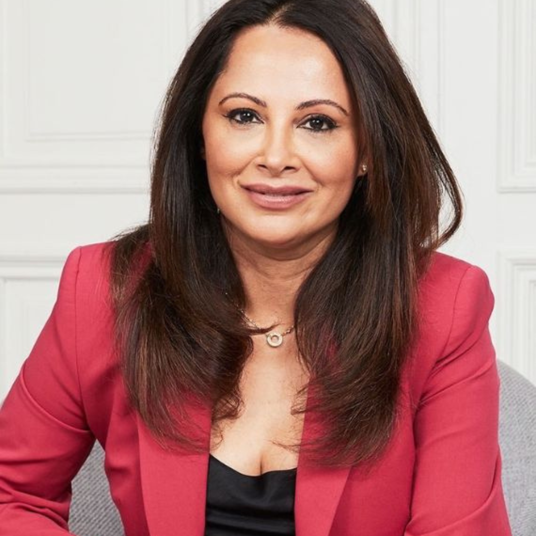 Smiling woman with long dark hair wearing a red blazer and black top seated against a white paneled wall.