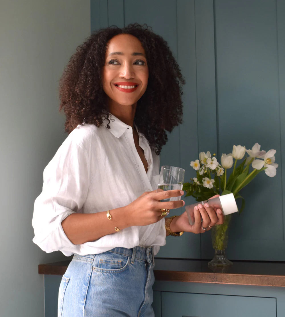 Woman holding a Lumity Morning Supplement bottle and a glass of water in a kitchen setting with flowers on the background