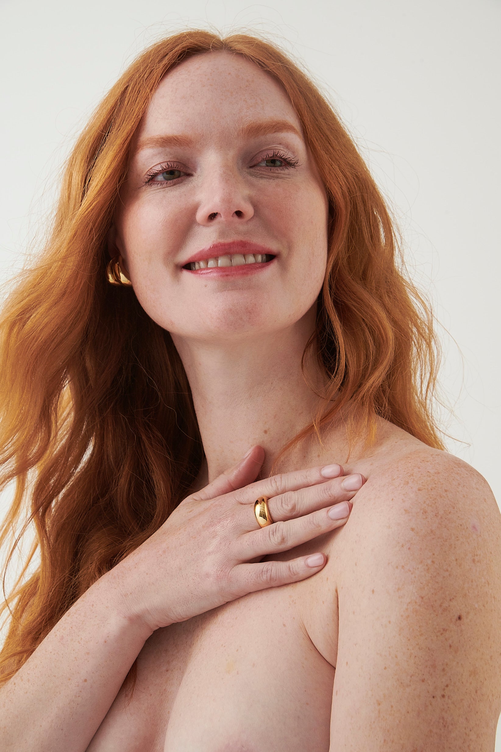 Smiling red-haired woman with clear skin gently touching her shoulder against a plain light background