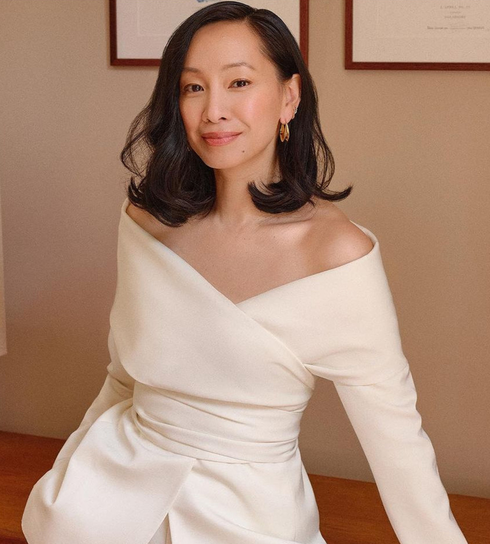 Woman with shoulder-length dark hair wearing an off-shoulder white top, smiling softly in a neutral-toned room.
