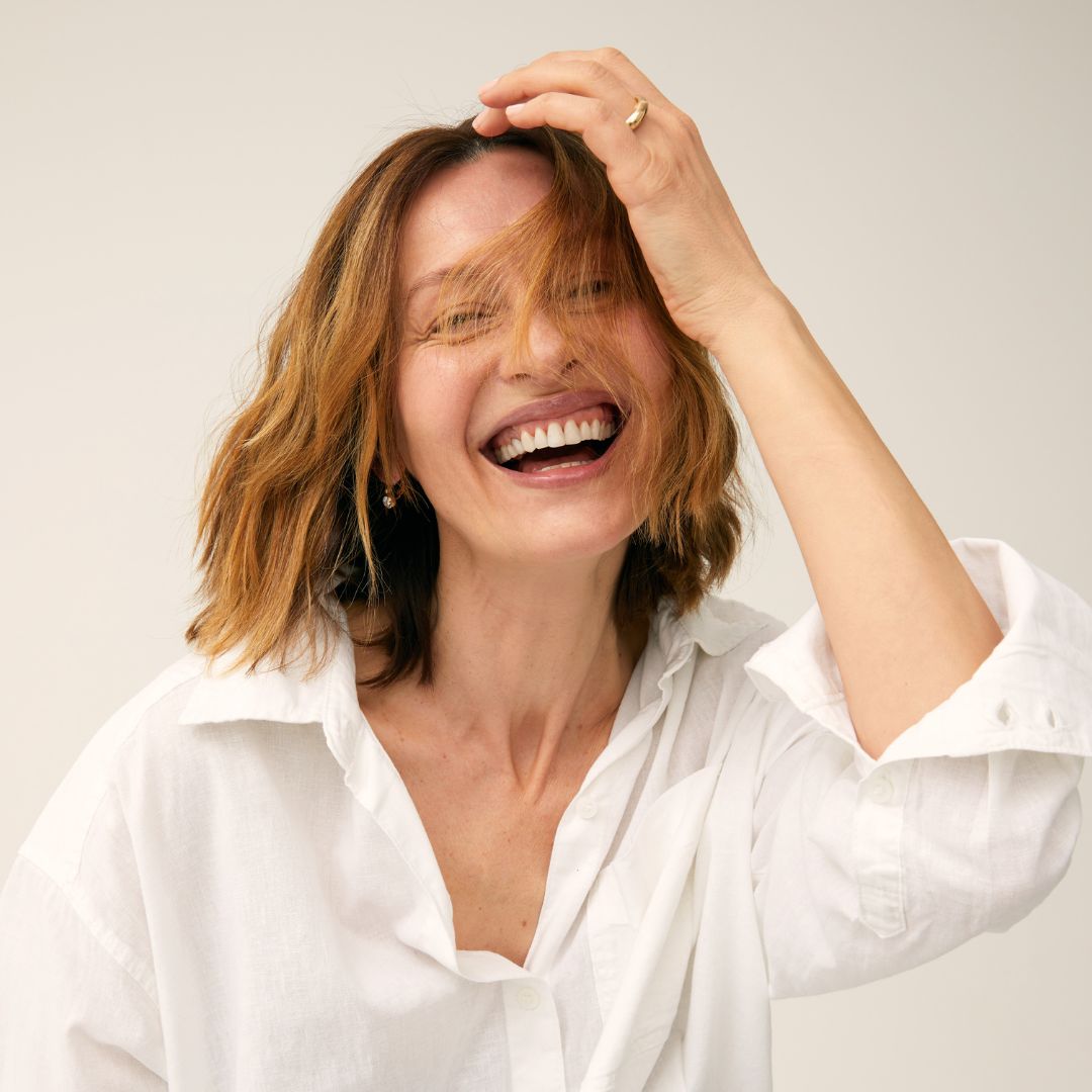 Woman in a white shirt laughing with one hand on her head against a plain background
