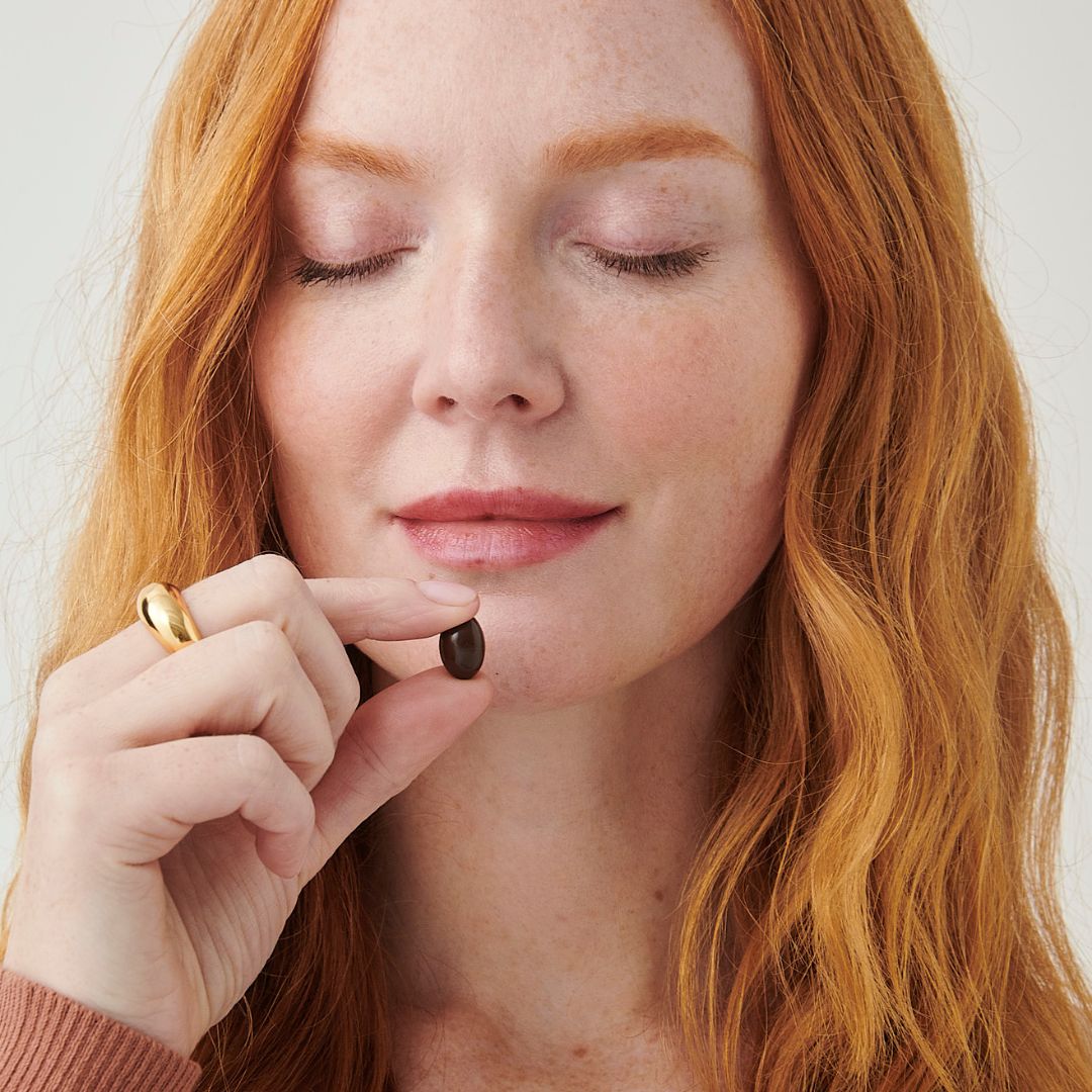 Woman with red hair holding a Lumity Night softgel her face against a neutral background