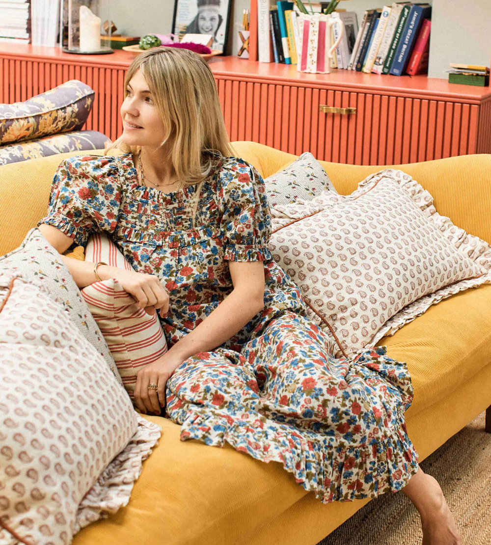 Matilda Goad  in floral dress sitting relaxed on a yellow couch surrounded by patterned pillows in a cozy living room