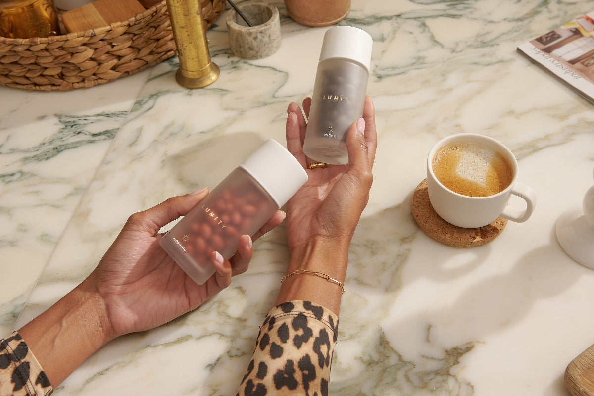 Hands holding Lumity Morning & Night for women on a marble surface with a cup of coffee in the background.